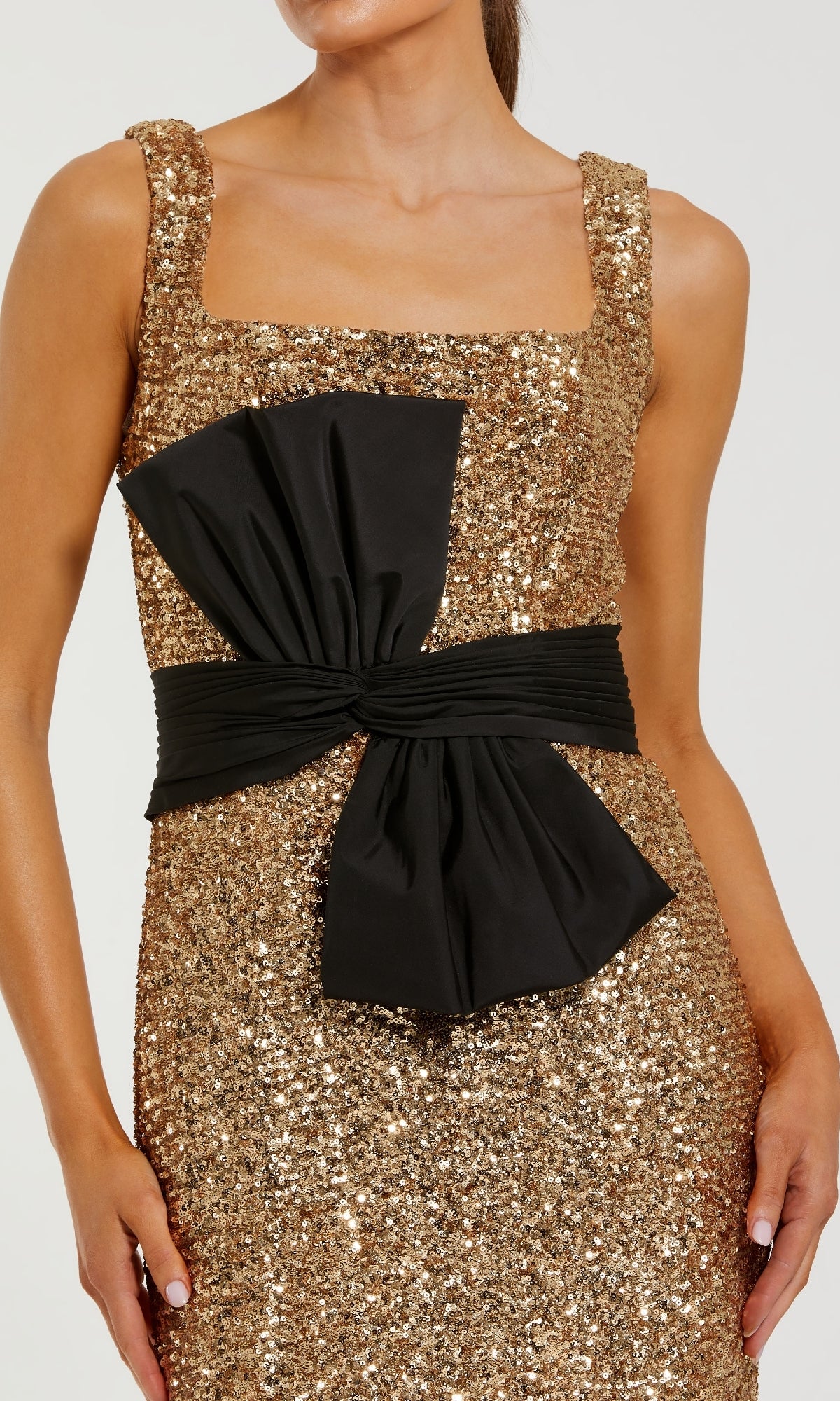 Close-up of a woman modeling a gold sequin dress with a black bow detail at the waist standing in front of a white background.